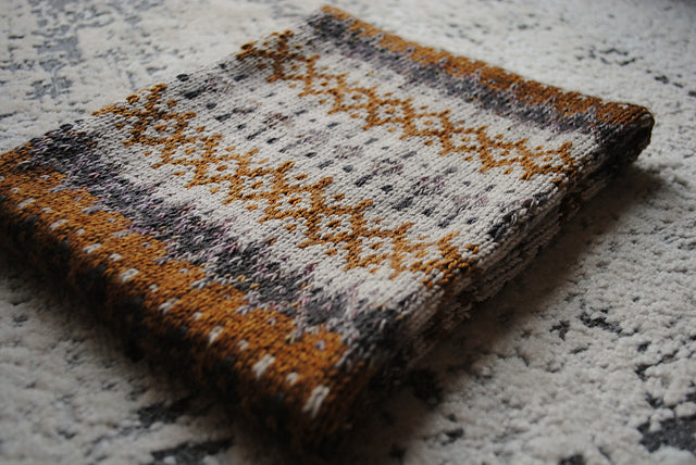 A folded up cowl on a speckled surface. The cowl features intricate colourwork details in shades of beige, grey, dark grey and orangey brown.