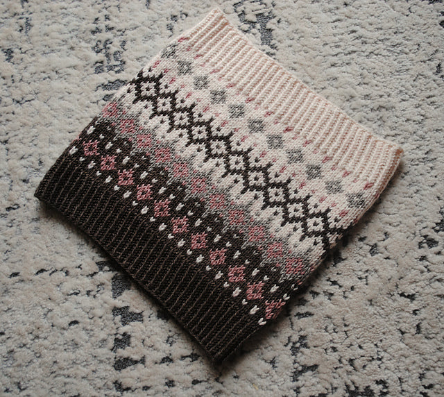 A narrow cowl on a speckled grey surface. The cowl features intricate colourwork detail in pinky beige, black, pink and purple.