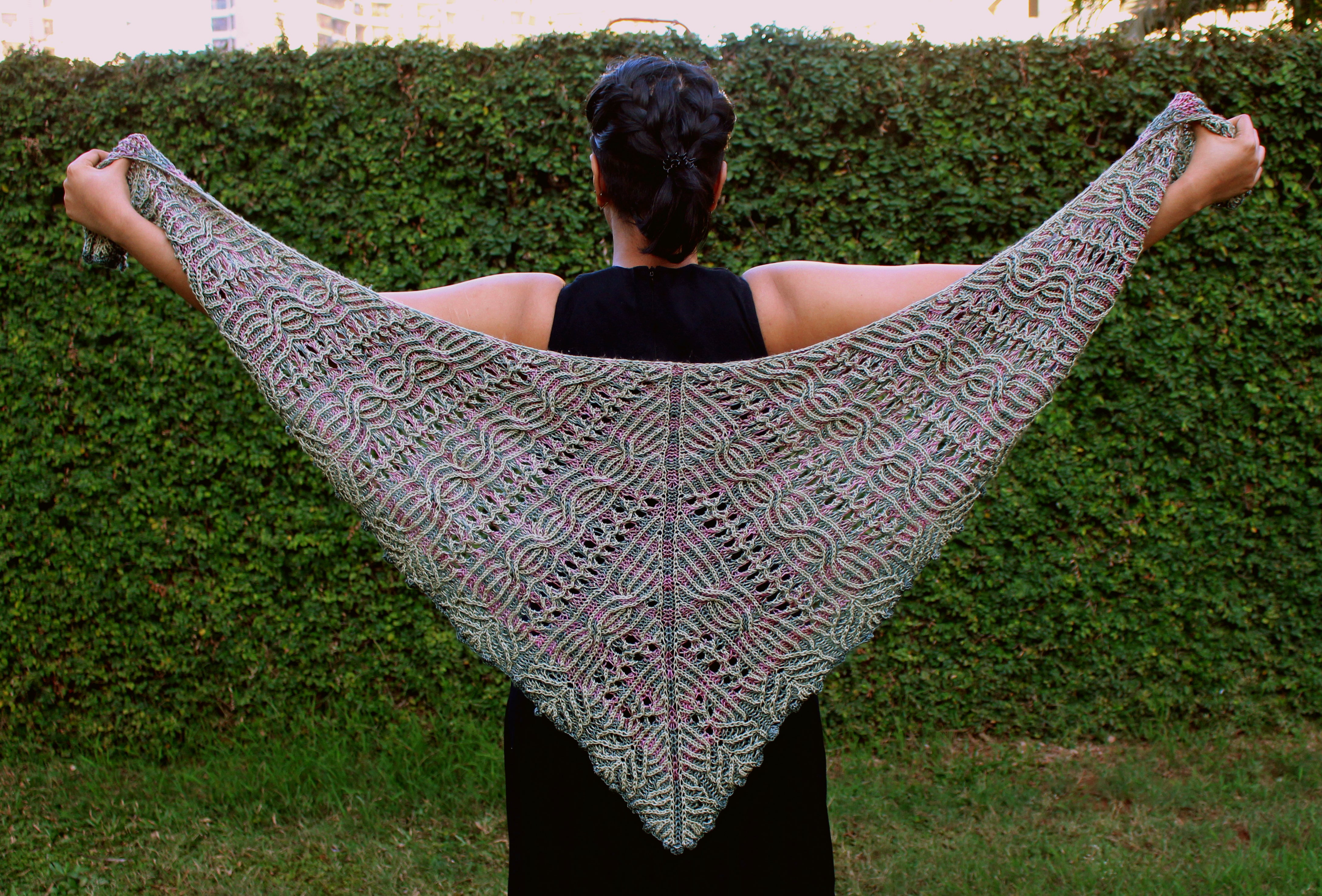 A person facing away from the camera towards a hedge. They are holding a large triangular shawl outstretched across their back. The shawl features intricate brioche lace patterns and has elements of pink and green to it. 