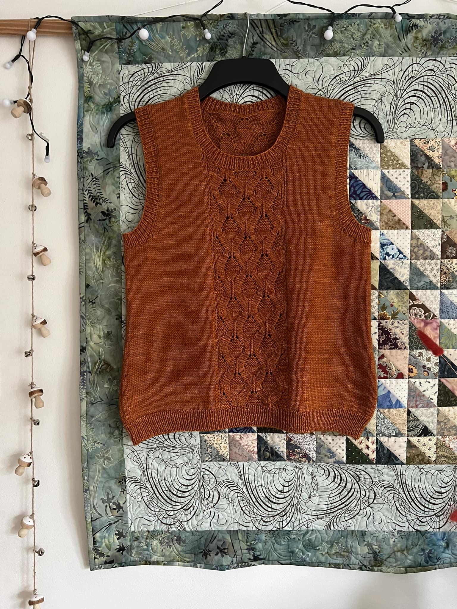 A rust coloured vest hanging in front of a patchwork quilt. The vest has a panel of cable details down the centre front