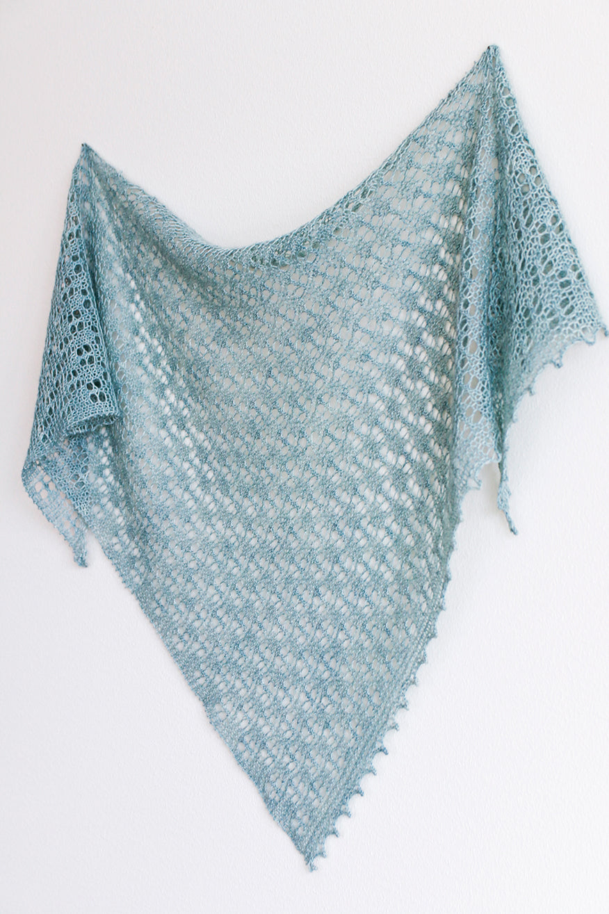 A blue triangular shawl hanging on a plain backdrop. The shawl has an all over simple lace pattern