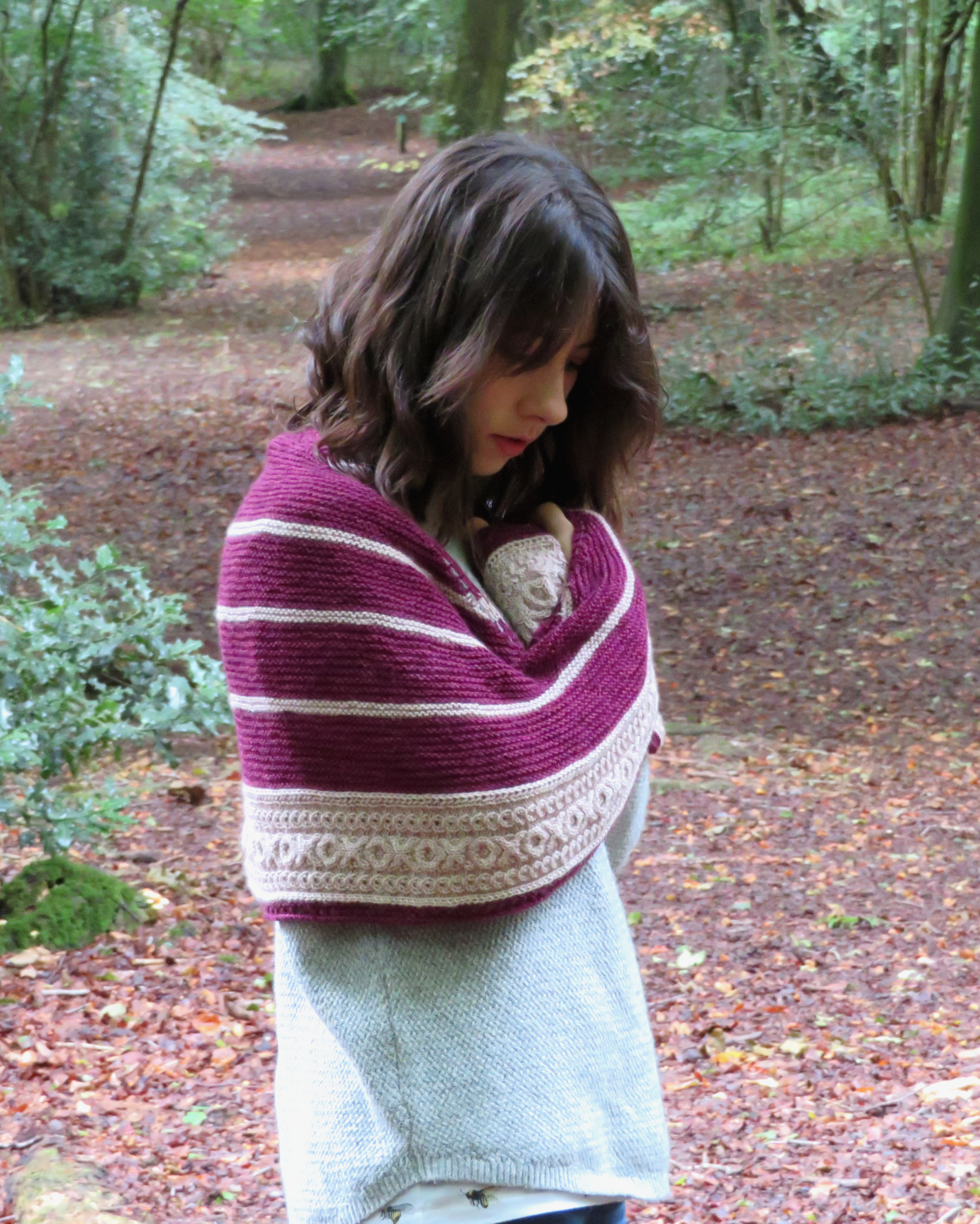 A person with a shawl wrapped around their shoulders. The shawl is crescent shaped with colourwork stripes of purple and beige