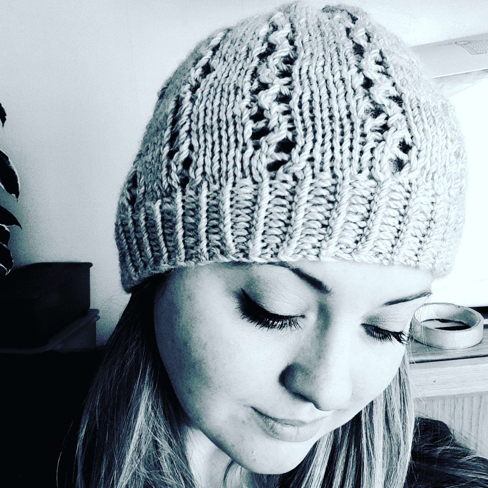 A black and white image of Hanna Gough looking towards the ground. Hanna is wearing a knitted beanie hat with textured lace details running the length of the hat.