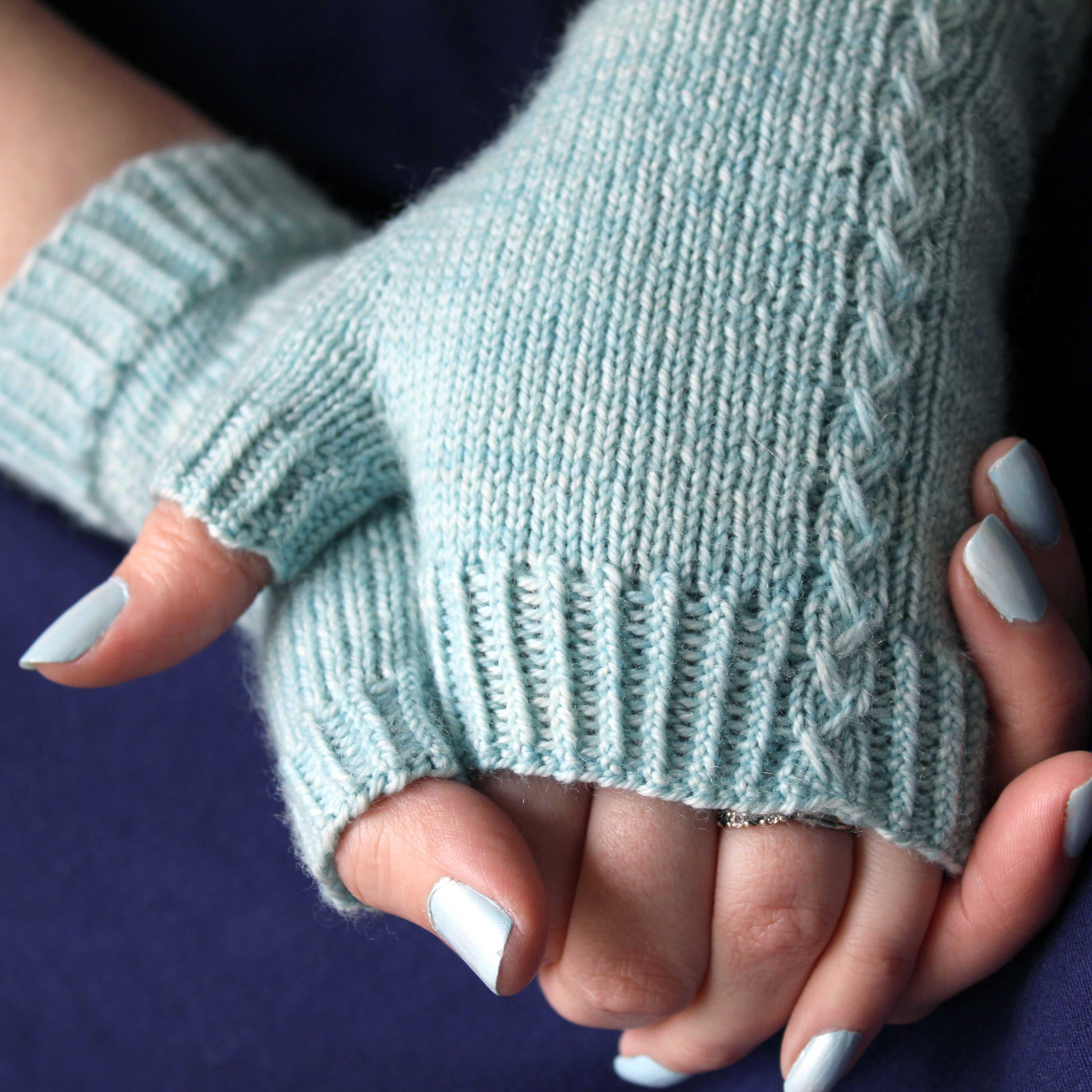 A pair of hands grasping each other wearing pale bright blue fingerless mitts. The mitts are mostly plain with a cable detail running up the outside of the hand