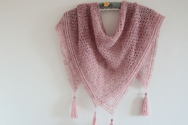 A pink lacey shawl draped over a hanger in front of a plain wall. The shawl features tassels on each point