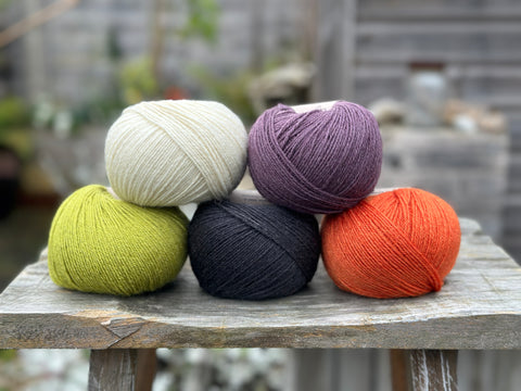 Five balls of yarn; the colours are cream, dark purple, lime green, charcoal grey, and rusty orange.