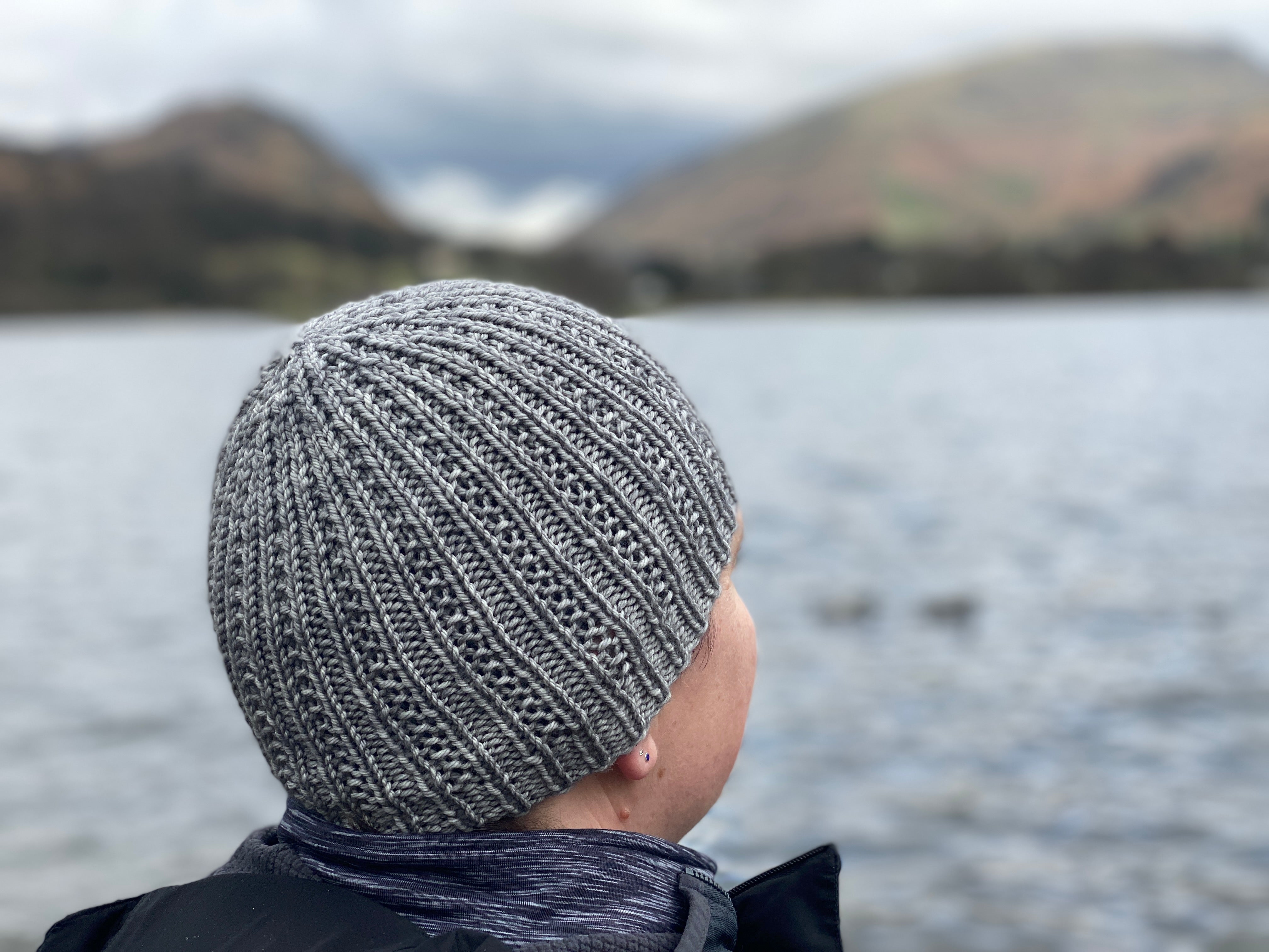 Victoria looking out over a lake towards mountains in the distance. She is wearing a grey knitted hat with textured stitch details running the height of the hat