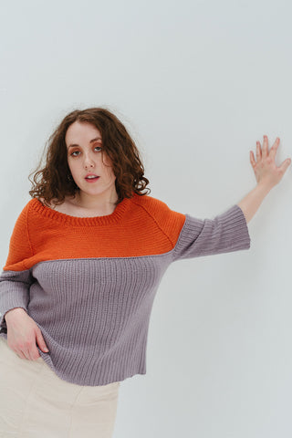 A person leaning one arm against a white wall. They are wearing a grey-purple and orange sweater