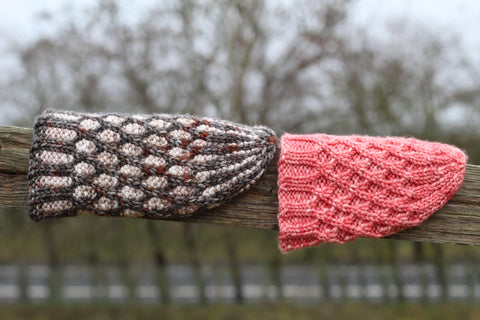 Two hats resting on a wooden fence. On the left hand side of the image is a two tone cable lattice in cream and beige with a dark grey and red contrast. On the right hand side is a bright pink hat in the same cable lattice design but one colour