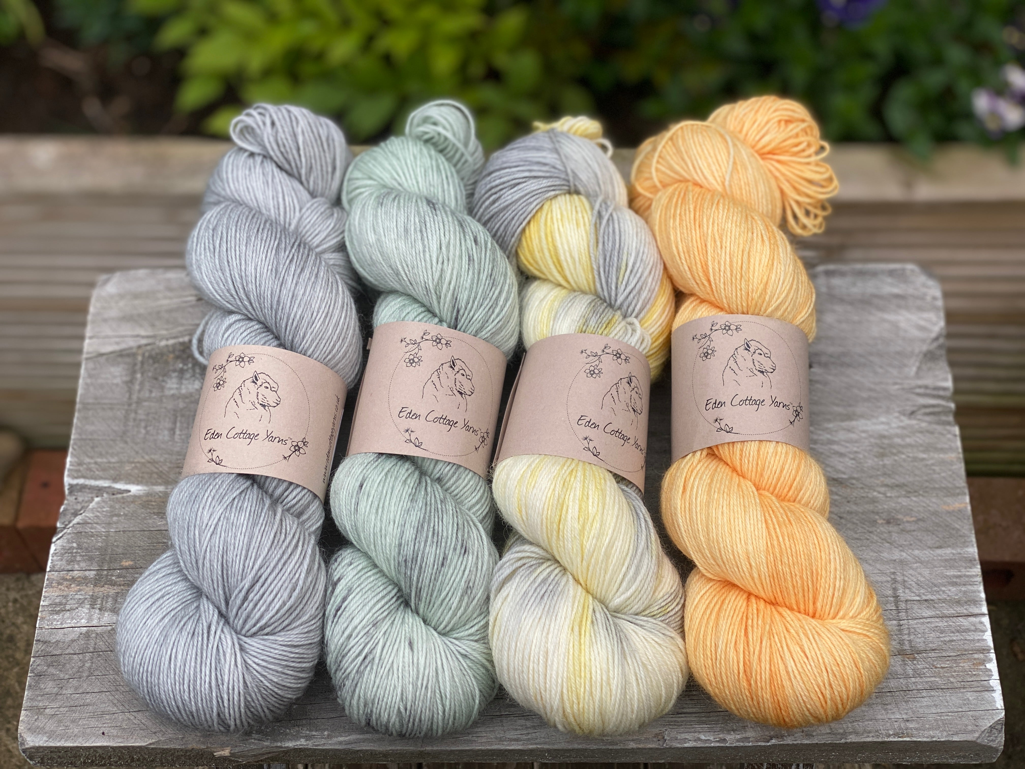 Four skeins of yarn. From left to right: a pale blue/grey yarn, a pale green yarn with grey splashes, a yellow, grey and cream yarn and an orange yarn