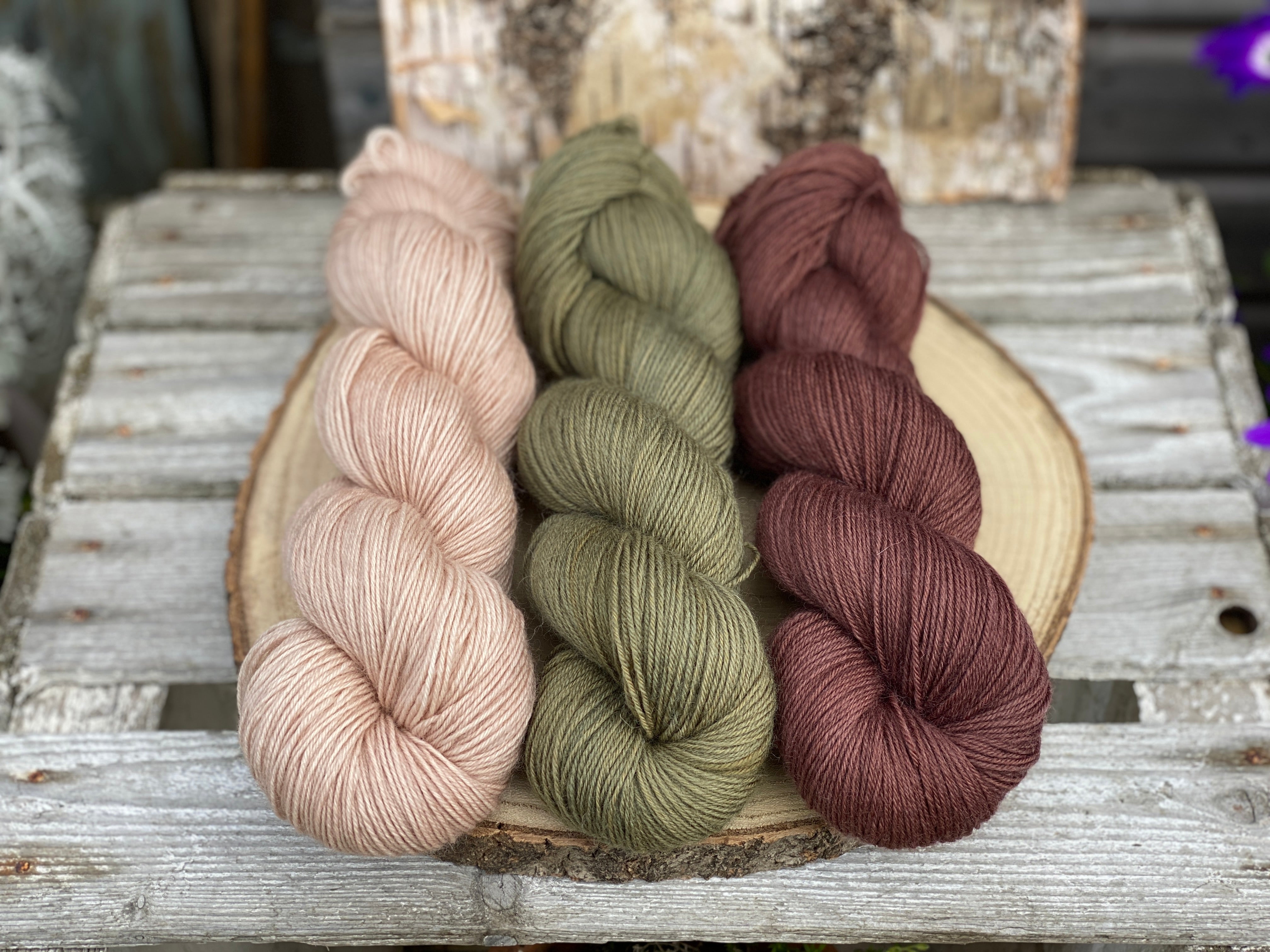 Three skeins of yarn. From left to right: a beige skein, a brownish green skein and a deep reddish brown yarn