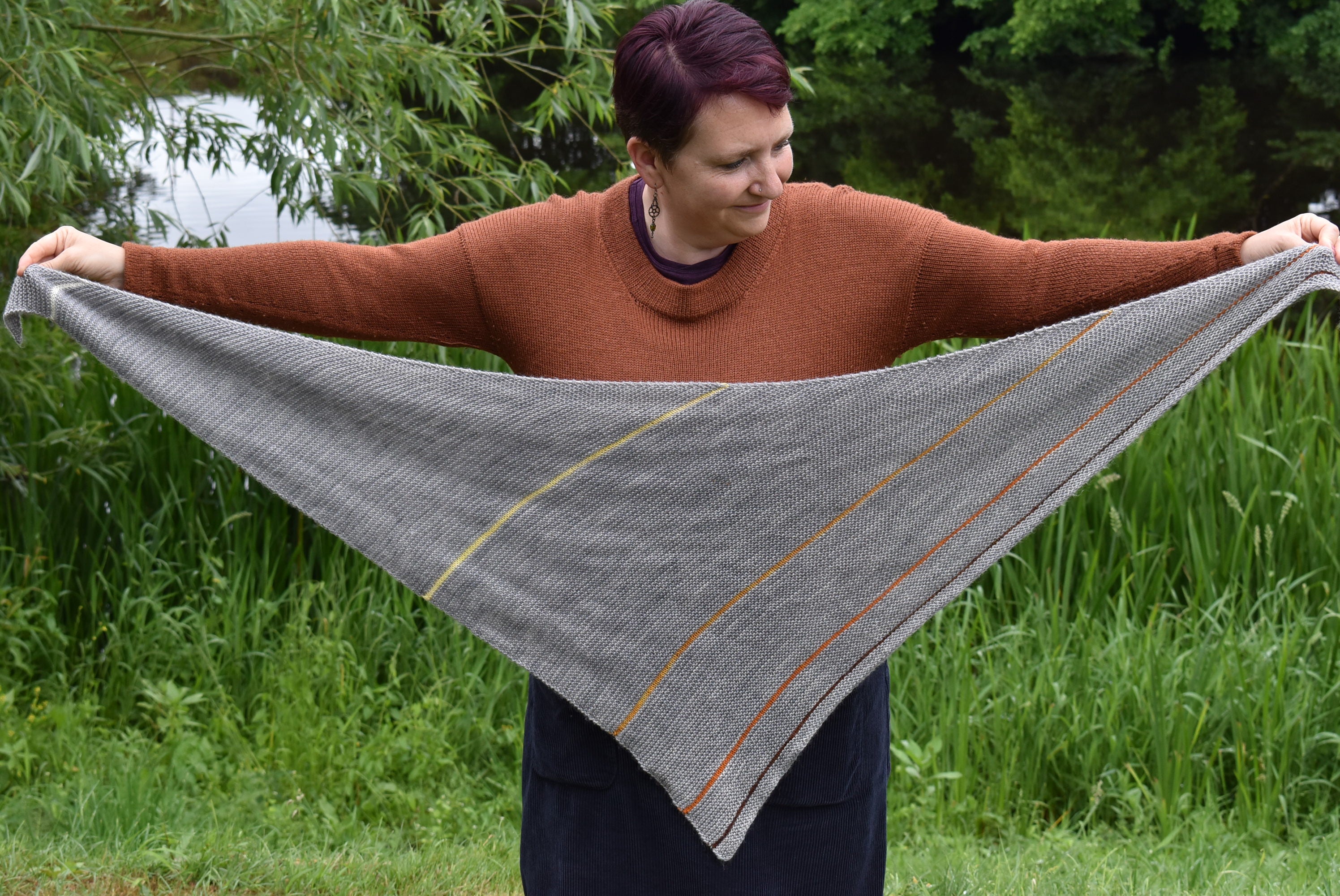 Victoria holding a grey triangular shawl outstretched. The shawl features small stripes in shades of yellow, orange and brown.