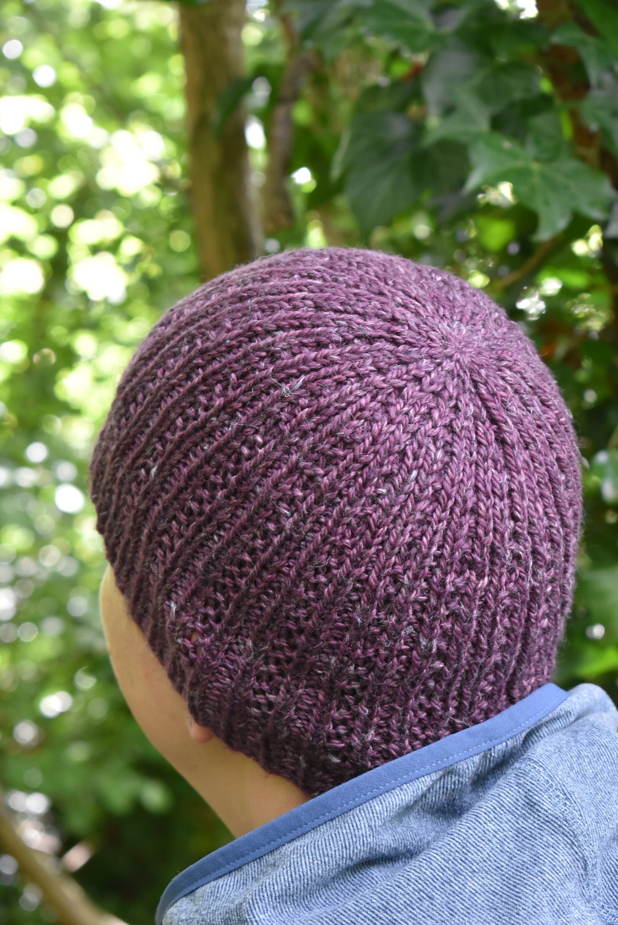 A person looking away from the camera towards woodland. They are wearing a purple knitted hat with a vertical stitch pattern.
