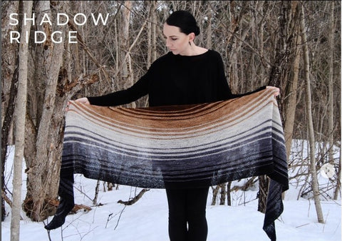 A person in a snowy landscape holding a semicircular shawl outstretched. The shawl fades through brown, cream and grey