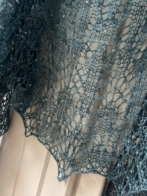 A delicate lace shawl in a shade of blue hanging against a white backdrop. The shawl has small beads throughout the design.
