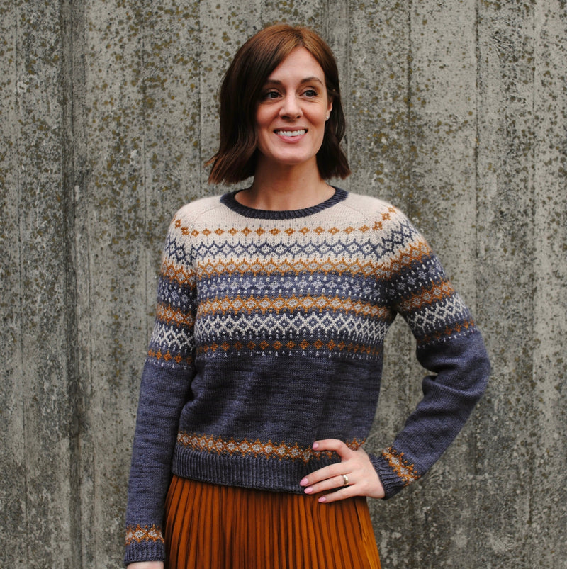 Rachel stood in front of a grey background wearing a detailed colourwork jumper in beige, grey, dark grey and a pop of orangey brown. 