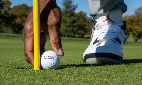 detail of a golf shoe and a hand picking up a golf ball from the green course