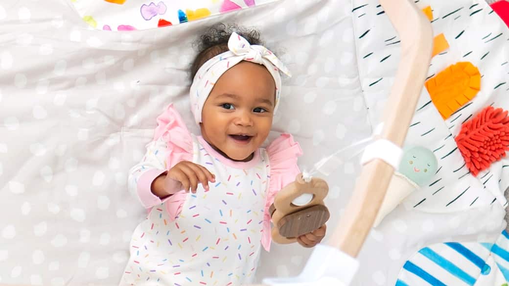 baby in rainbow sprinkles print holding toy from play yard