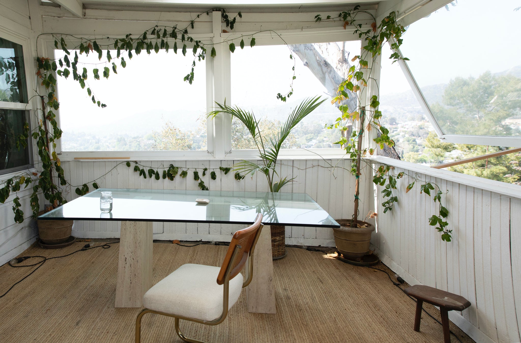A photo of Maggi Simpkins scared summertime space: a porch that has been converted into a sunroom, filled with plants and a large glass desk.  