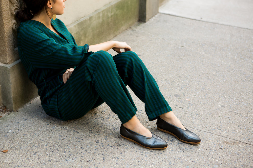 Image of a model seated wearing the Coclico Polaris flat that has been styled with a simple jumpsuit. 