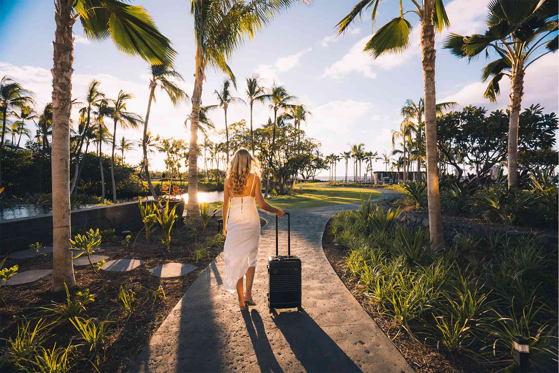 A girl in white dress with black Arlo Skye Frame Carry-On Max walking down a pathway in a lush tropical garden.