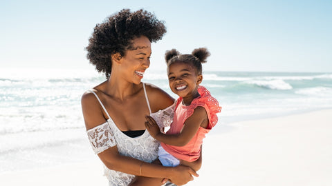 Woman carrying her young daughter, both smiling at the beach