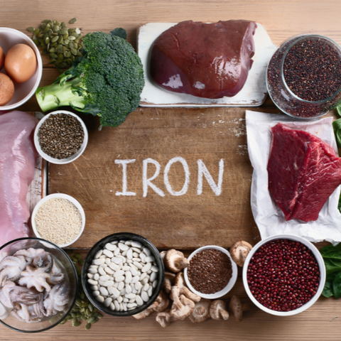 A wooden board with a text "Iron" surrounded by different red meat, seeds and broccolli
