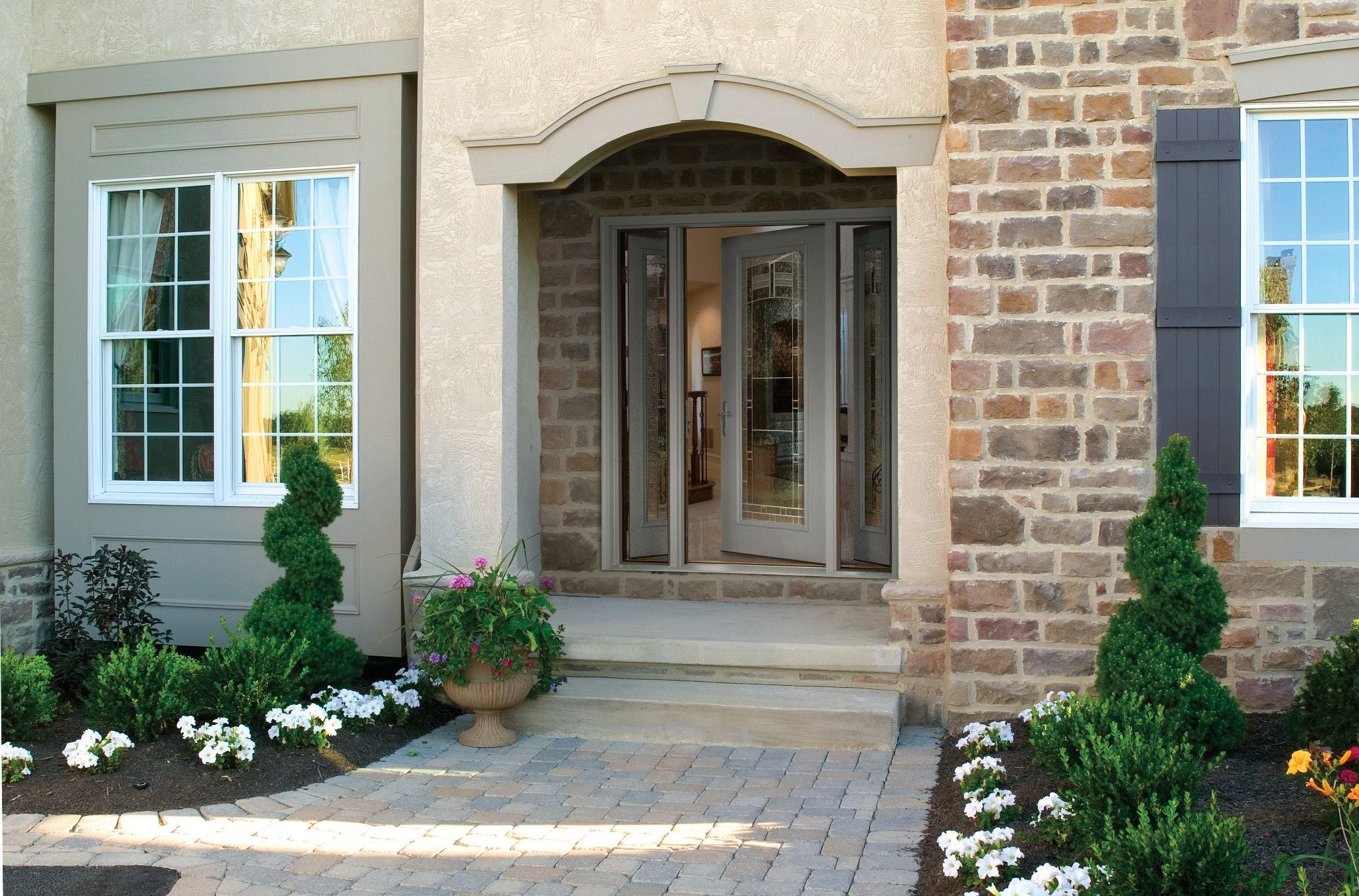 This house has a stone walkway and a covered entrance decorated by a PVC trim header . There are scrubs and flowers in the flowerbeds next to the walkway. 