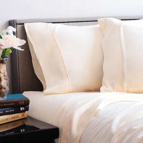 Elegant ivory bed sheets and pillowcases set on a stylish bed with a dark headboard, complemented by a bedside stack of books and a vase with pink flowers, presenting a clean and inviting bedroom atmosphere