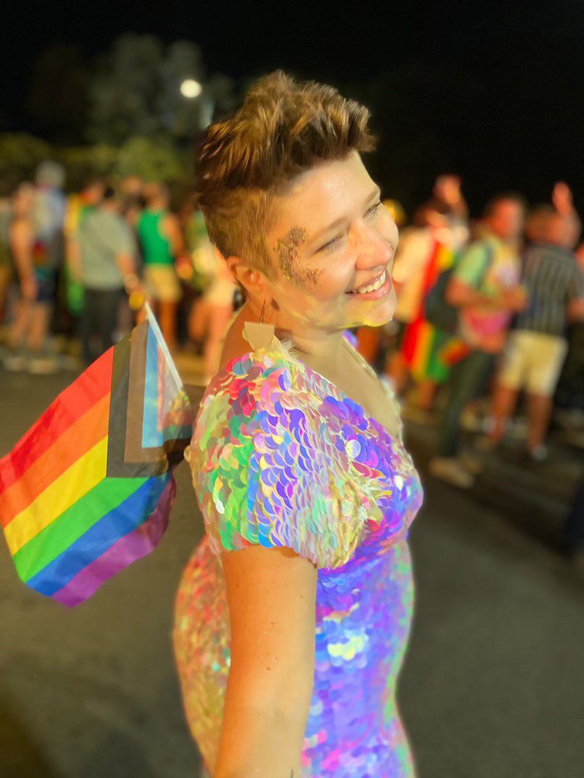 Smiling woman with short styled hair at Sydney Gay Pride holding a pride flag and wearing a Rosa Bloom Aphrodite Sequin Jumpsuit in Opal white colour