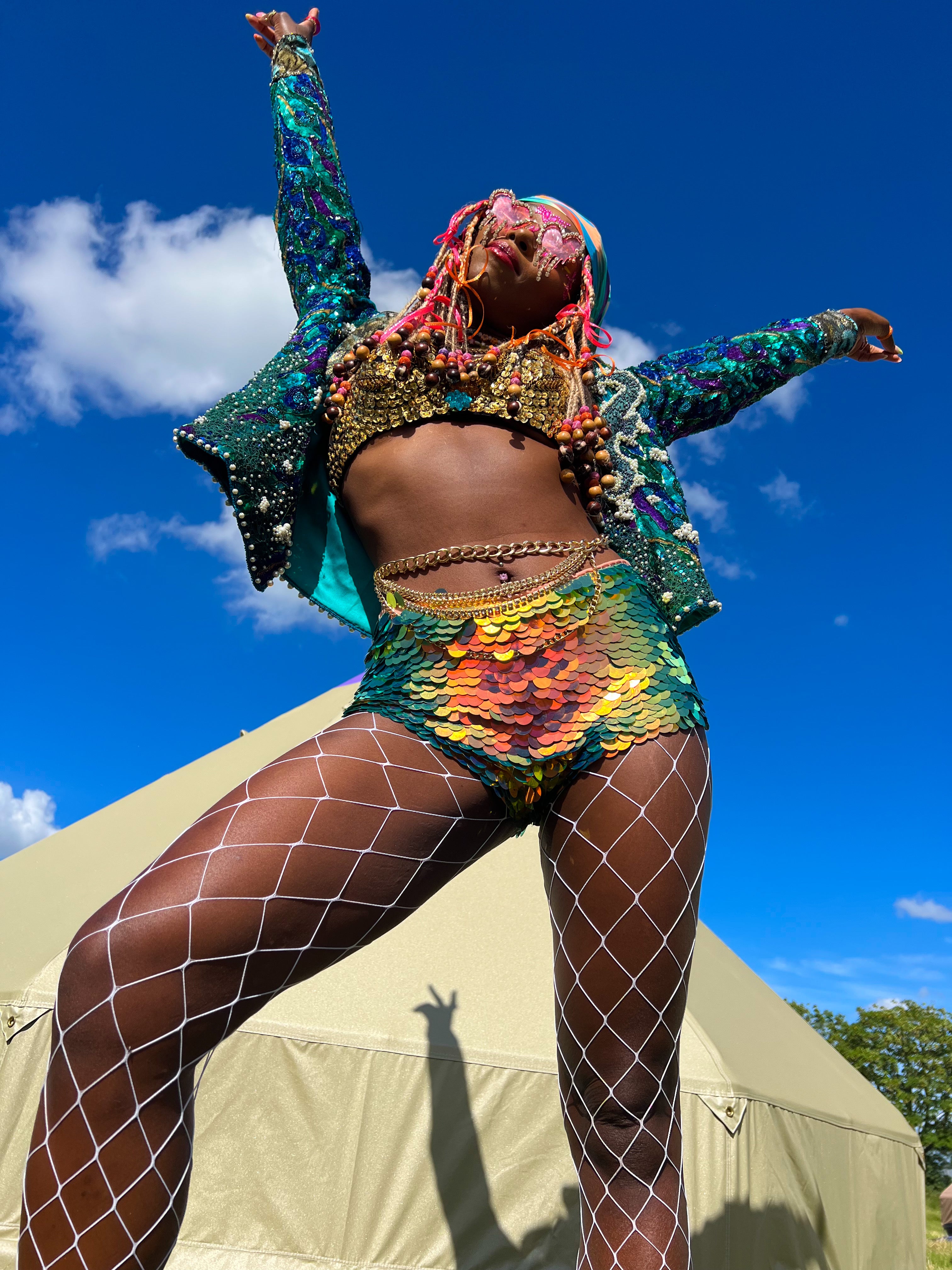 Up facing view of woman posing with her arms in the blue sky in front of a tipi tent wearing sparkly festival outfit including Rosa Bloom sequin hot pants