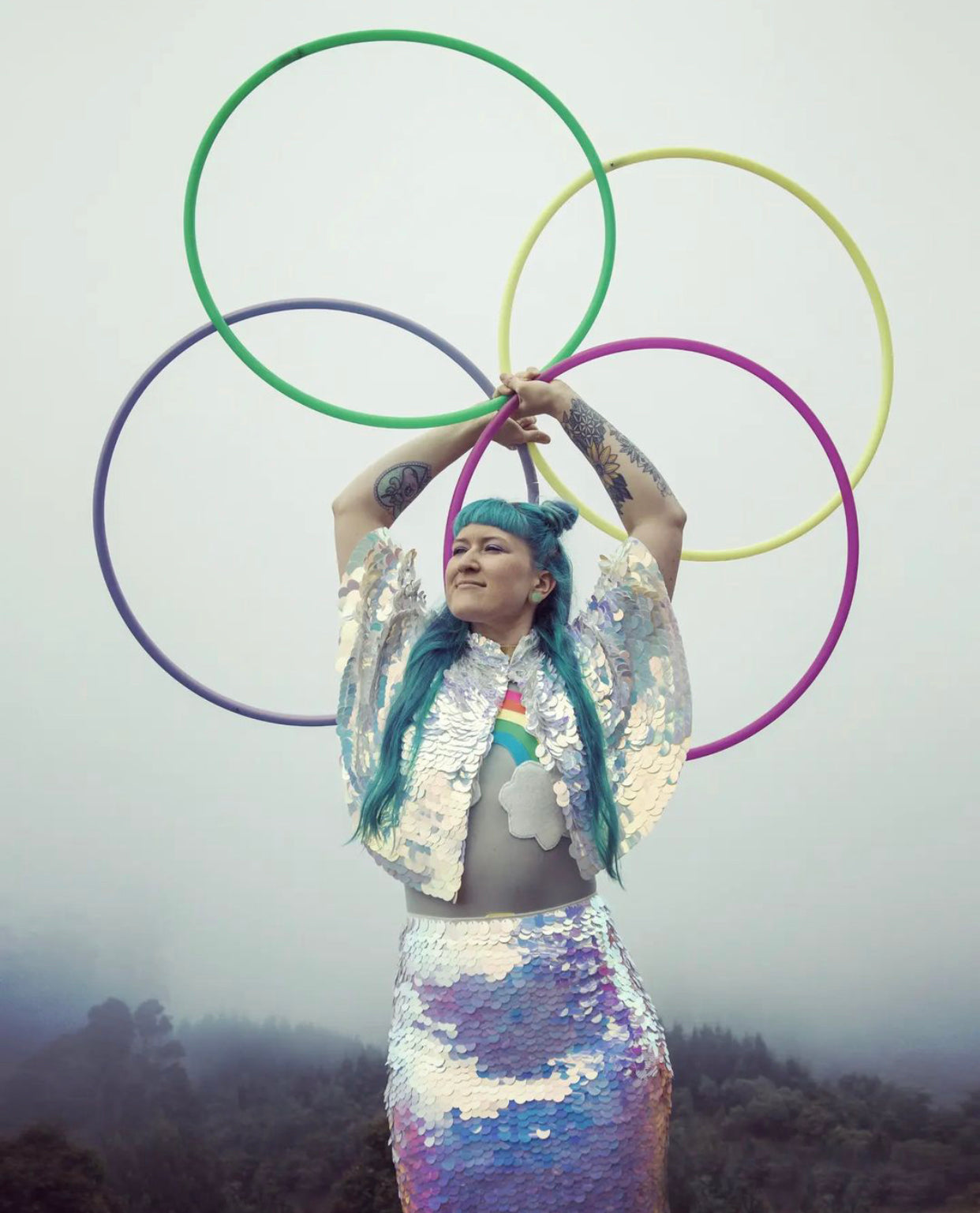Blue haired woman in Rosa Bloom Opal sequin cape and skirt posing with mutilcoloured hula hoops with a misty forest backdrop