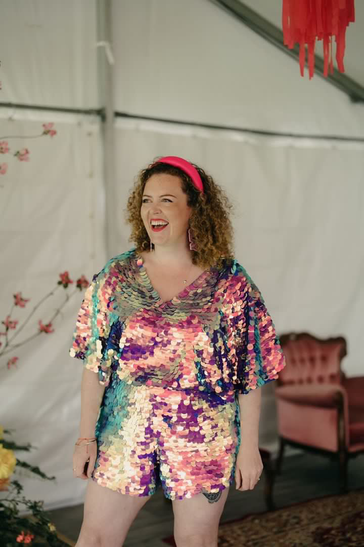 Laughing woman with curly hair wearing Rosa Bloom Sequin Cape Playsuit, the Mella in a purple pink orchid colour