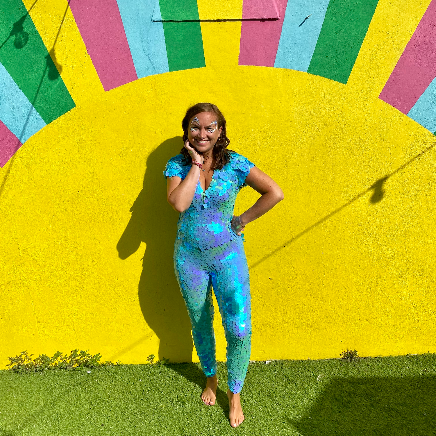 Smiling petite lady posing in front of a painted sun wall wearing a Rosa Bloom Aphrodite Sequin Jumpsuit in Mint colour