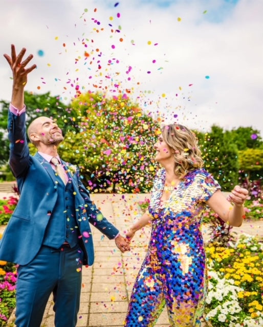 A bride wearing a purple sequin jumpsuit stands under a cloud of colourful confetti with her husband.