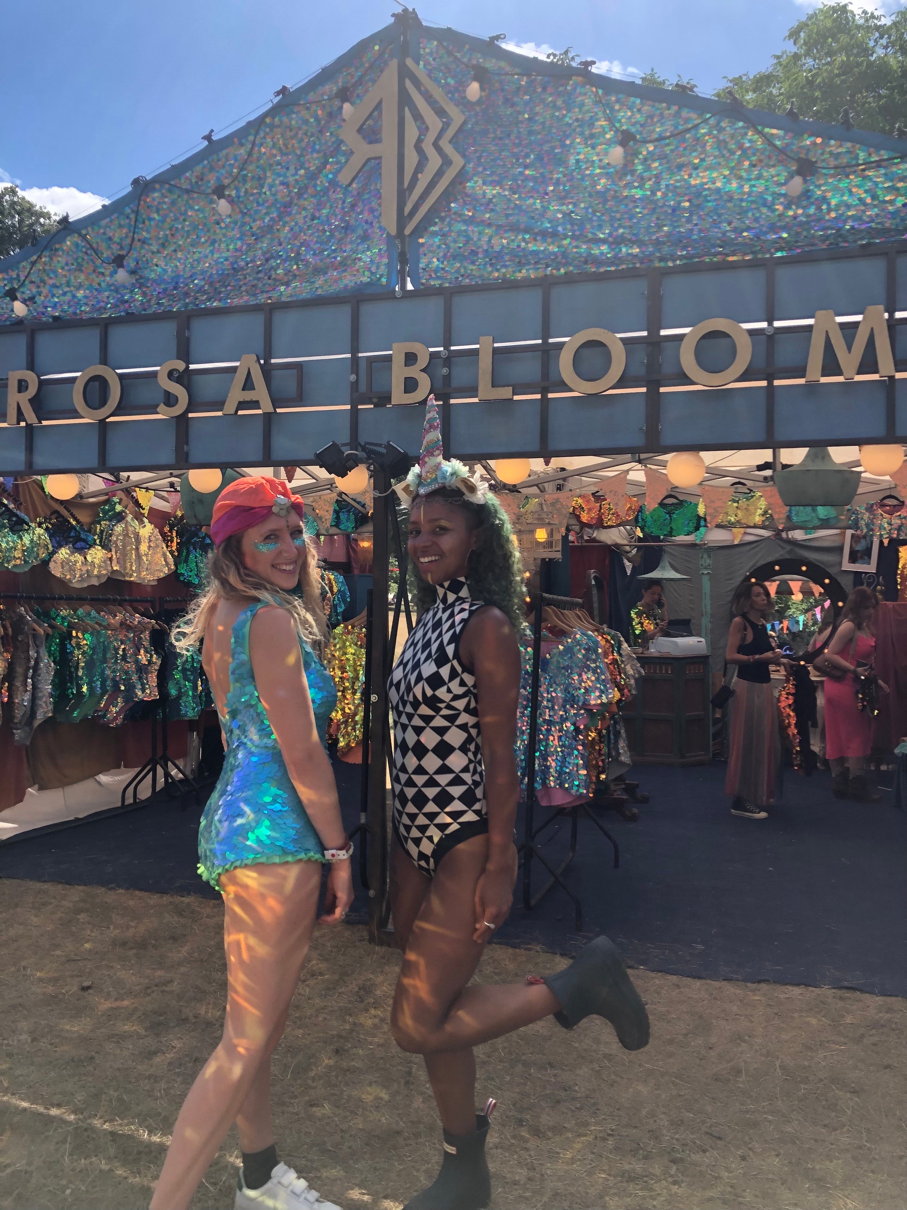Two smiling young women on a summer's day stood in front of the Rosa Bloom stall at Wilderness Festival wearing the Sea Circus playsuit in Mint and the Fifi leotard in Tri Print