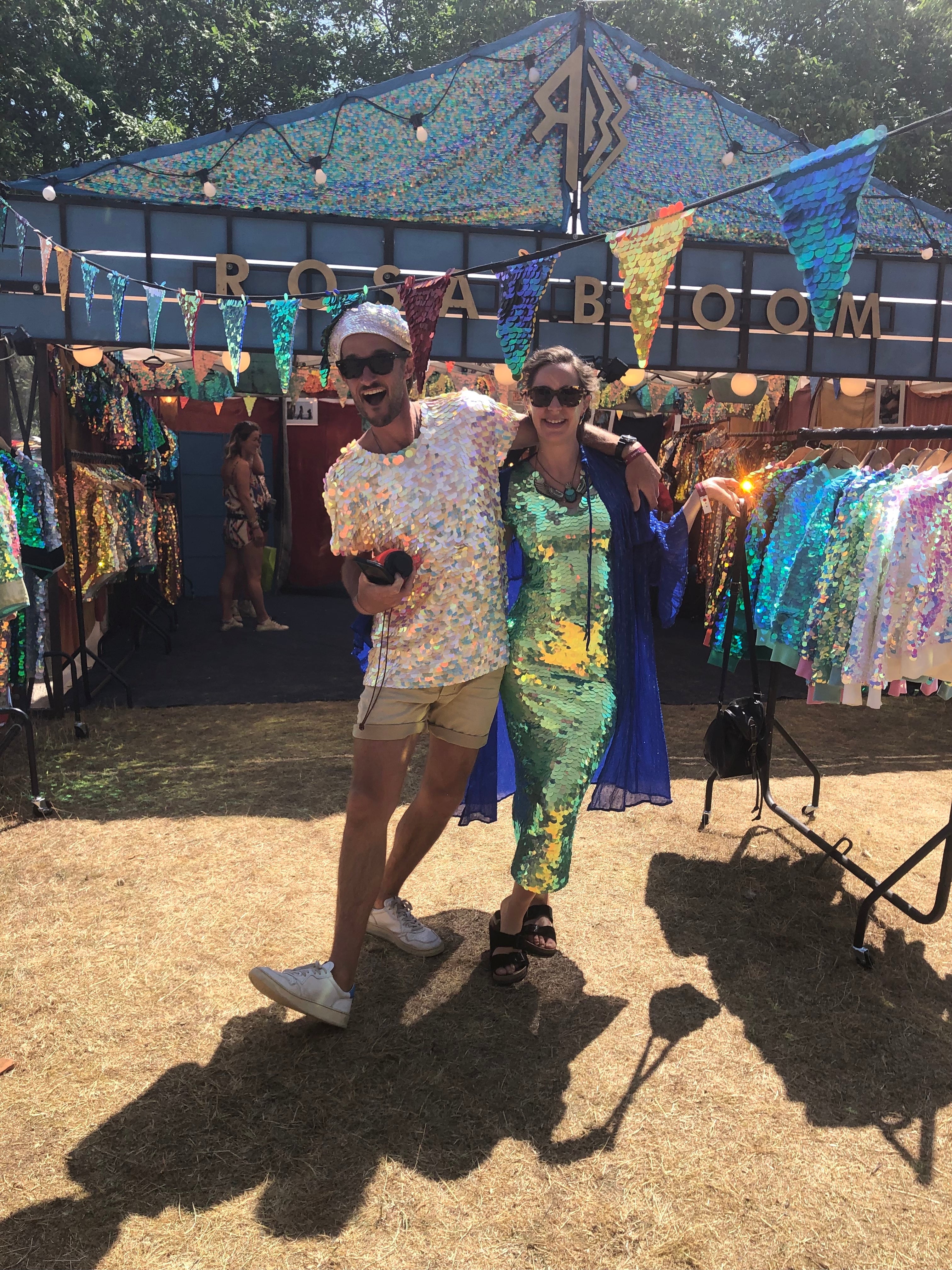 Smiling couple wearing black sunglasses standing in front of the Rosa Bloom stall at Wilderness festival wearing the Rosa Bloom Georgie dress in Chameleon and the Grace Sequin Beret in Opal