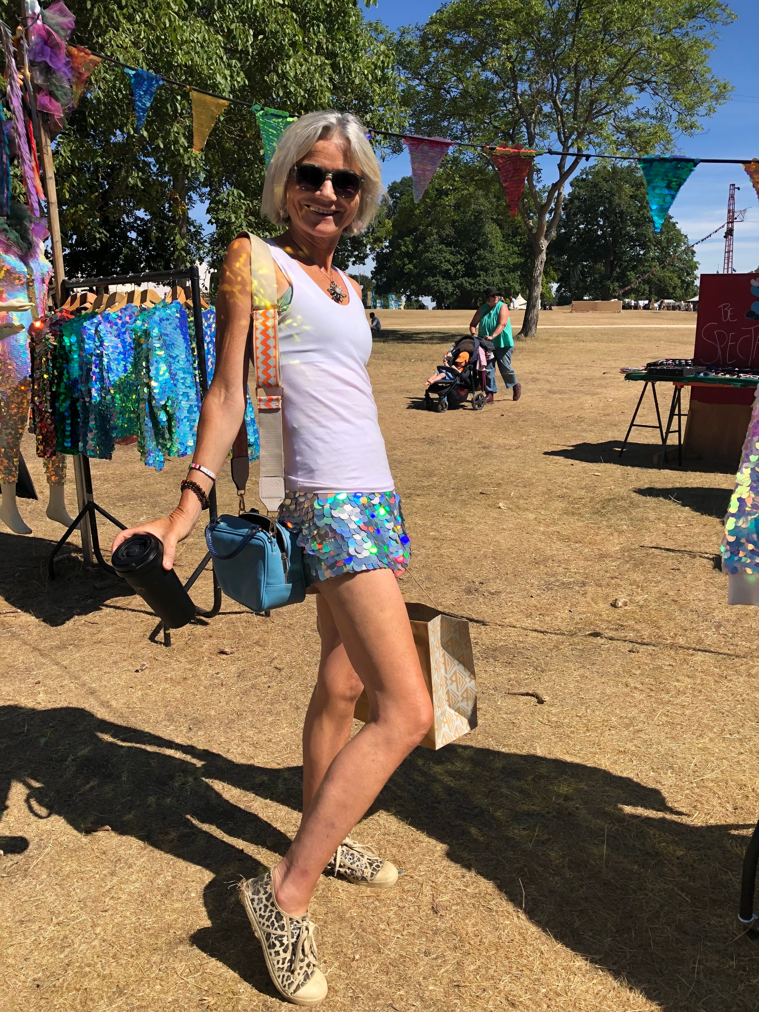 Smiling woman wearing black sunglasses and blonde bob stood in front of sequin bunting at the Rosa Bloom stall at Wilderness Festival wearing the Juno Sequin shorts in Hologram Silver