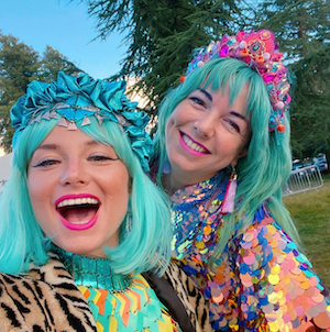 Two friends in blue wigs and sequin outfits smile as thet pose for a selfie photo.