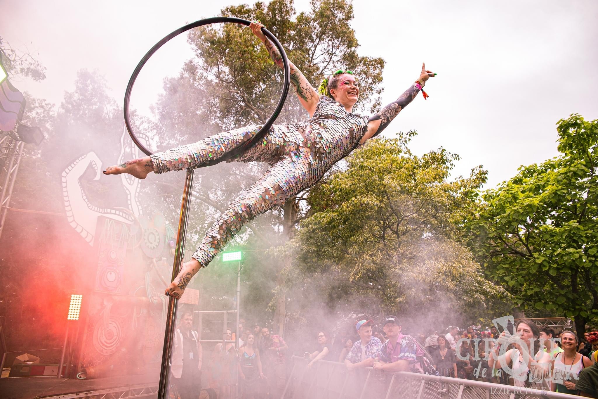 Circus performer balancing on an aerial hoop on a stage surrounded by trees, smoke and red lights wearing a holographic silver sequin Rosa Bloom Jumpsuit at Isle of Wight Festival