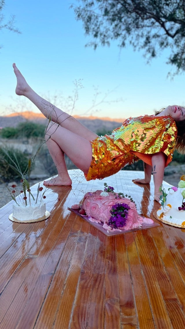 Joyful woman at sunset in bridge pose on top of a varnish oak table smiling above a pink cake wearing a Rosa Bloom Mella jumpsuit in Coral