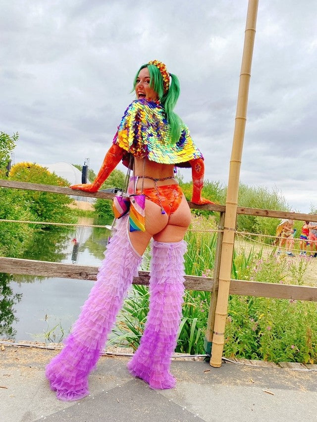 Girl with green hair stood on a bridge over a stream wearing a multicoloured festival outfit. including assless chaps and a multi coloured Rosa Bloom Eclipse sequin cape