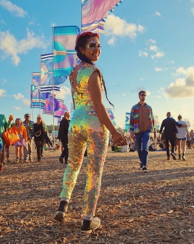 Girl looking over her shoulder at Glastonbury Festival stood in front of colourful flags wearing a Rosa Bloom sequin Aphrodite jumpsuit