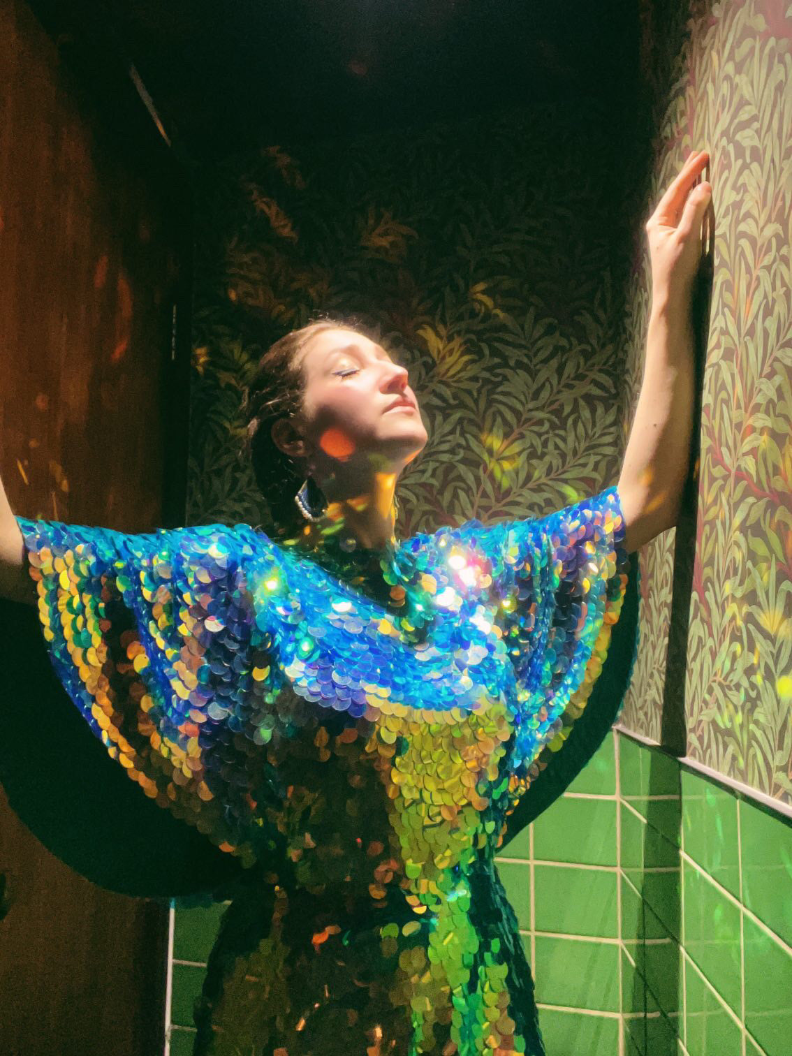 Woman in sequin romper by Rosa Bloom poses in green tiled bathroom