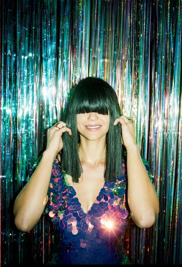 Laura Leezy smiling with her fringe covering her eyes in front of a sparkly show curtain wearing a Rosa Bloom Sequin Aphrodite Jumpsuit