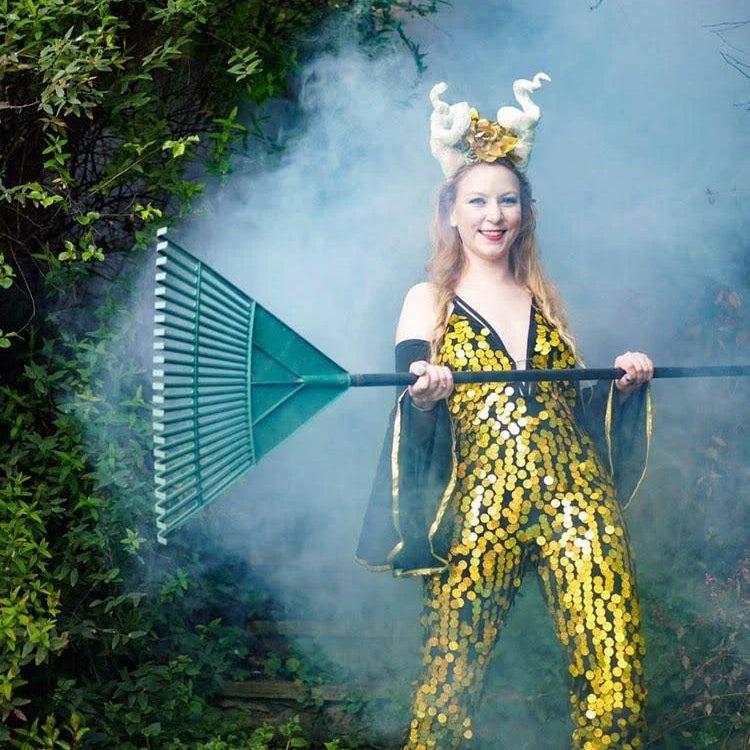 Woman holding garden rake wears a horn headpiece and yello sequin jumpsuit