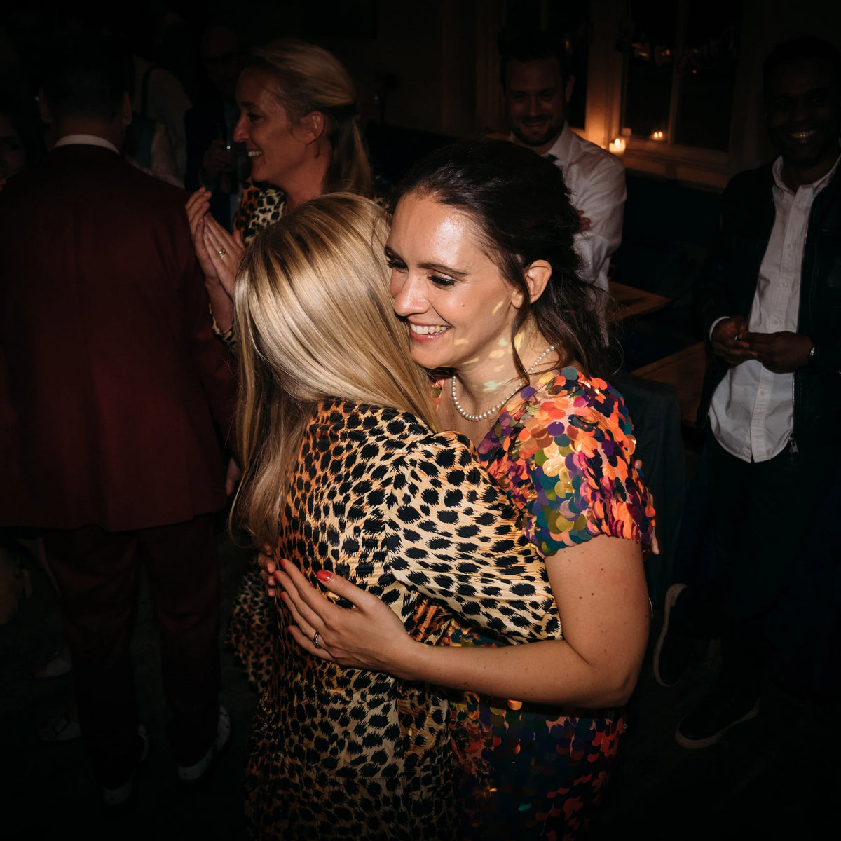 A bride wearing an orange sequin jumpsuit embraces her bridesmaid who is wearing a leopard print dress.