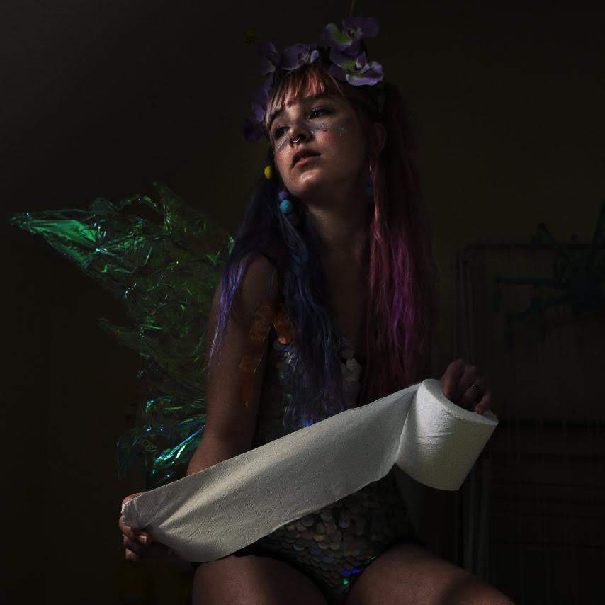 Woman with fairy wings and a sequin leotard poses in a dark room with toilet roll