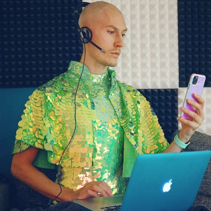 Man in a lime green cape and leotard sitting in bead with a laptop and headset 