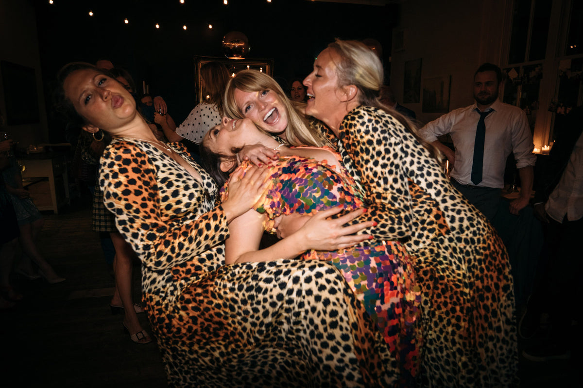 A bride wearing a festival style sequin jumpsuit is being embraced by several of her bridesmaids who are all wearing leopard print. 